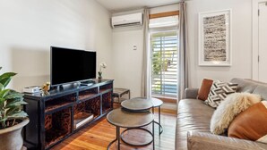 A modern living room with a flat-screen TV on a wooden console, a grey sofa with patterned cushions, a small round table, and a potted plant. - The Driftwood on Magazine (New Orleans)