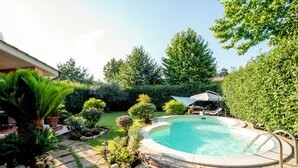A circular swimming pool with a stone deck surrounding it, a patio area with outdoor furniture, and a variety of landscaped greenery including bushes and trees. - The Aurora's Nest (Belvedere Lidia)