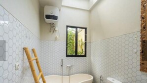 Un baño con una bañera independiente, inodoro y ventana con vistas a la vegetación.