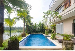 Una piscina rectangular rodeada de plantas tropicales y un edificio moderno con balcones.
