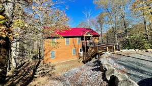 Un chalet rustique avec un toit rouge, une terrasse en bois et un chemin de pierre menant à l’entrée.