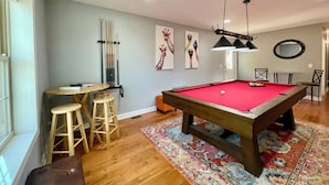 Une table de billard recouverte d’un feutre rouge, des tabourets en bois, un porte-queues et une table de billard.