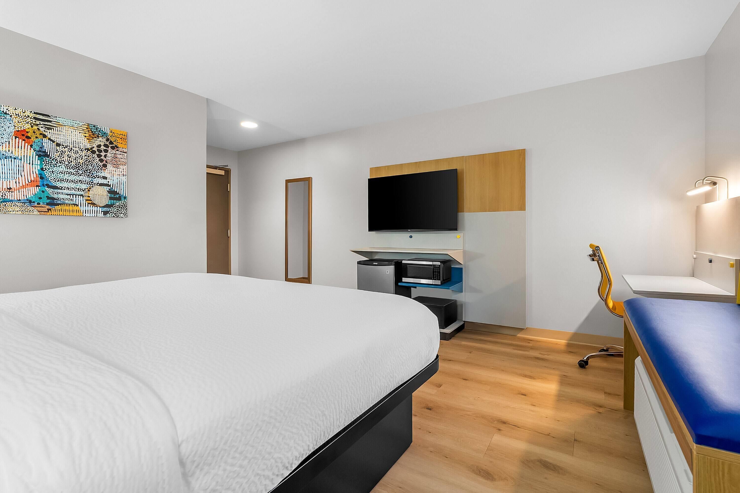 A modern hotel room with a large bed, a flat-screen TV mounted on the wall, a small desk with a computer, and a guitar leaning against the wall.