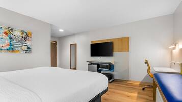 A modern hotel room with a large bed, a flat-screen TV mounted on the wall, a small desk with a computer, and a guitar leaning against the wall.