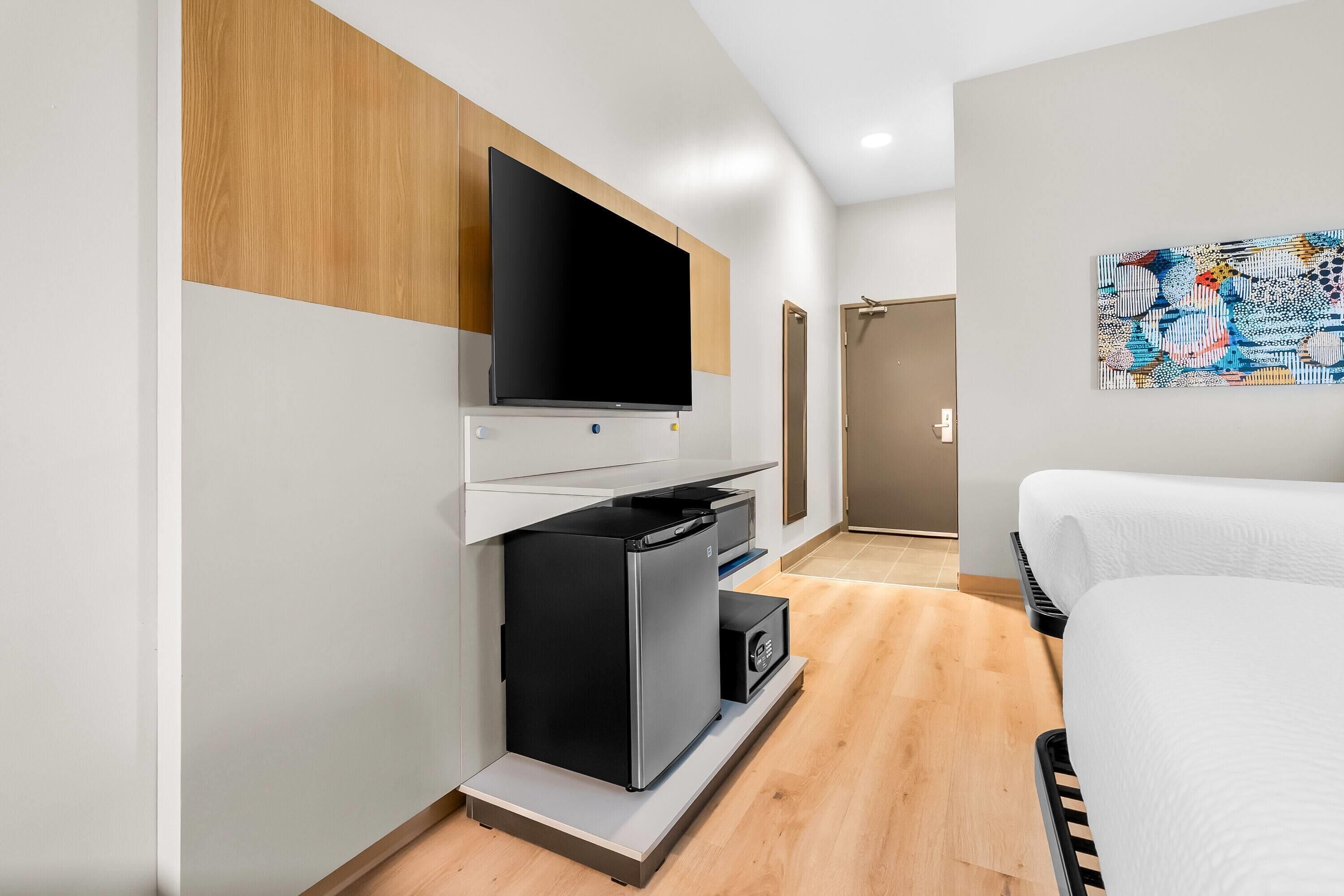 Een moderne hotelkamer met een flatscreen-tv bevestigd aan een houten paneel, een kleine keukenunit, een witte bank en een kleurrijk abstract schilderij aan de muur.