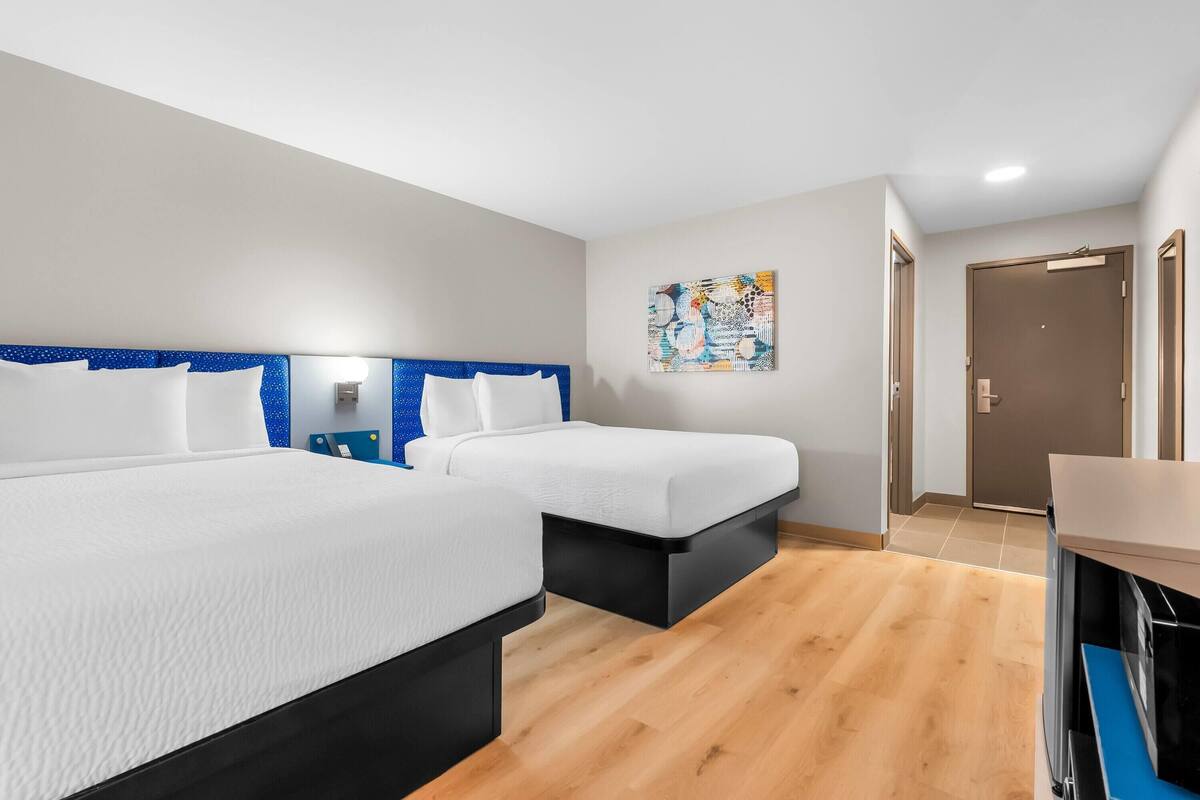 Quarto de hotel com duas camas, piso de madeira, um quadro na parede e uma porta.
