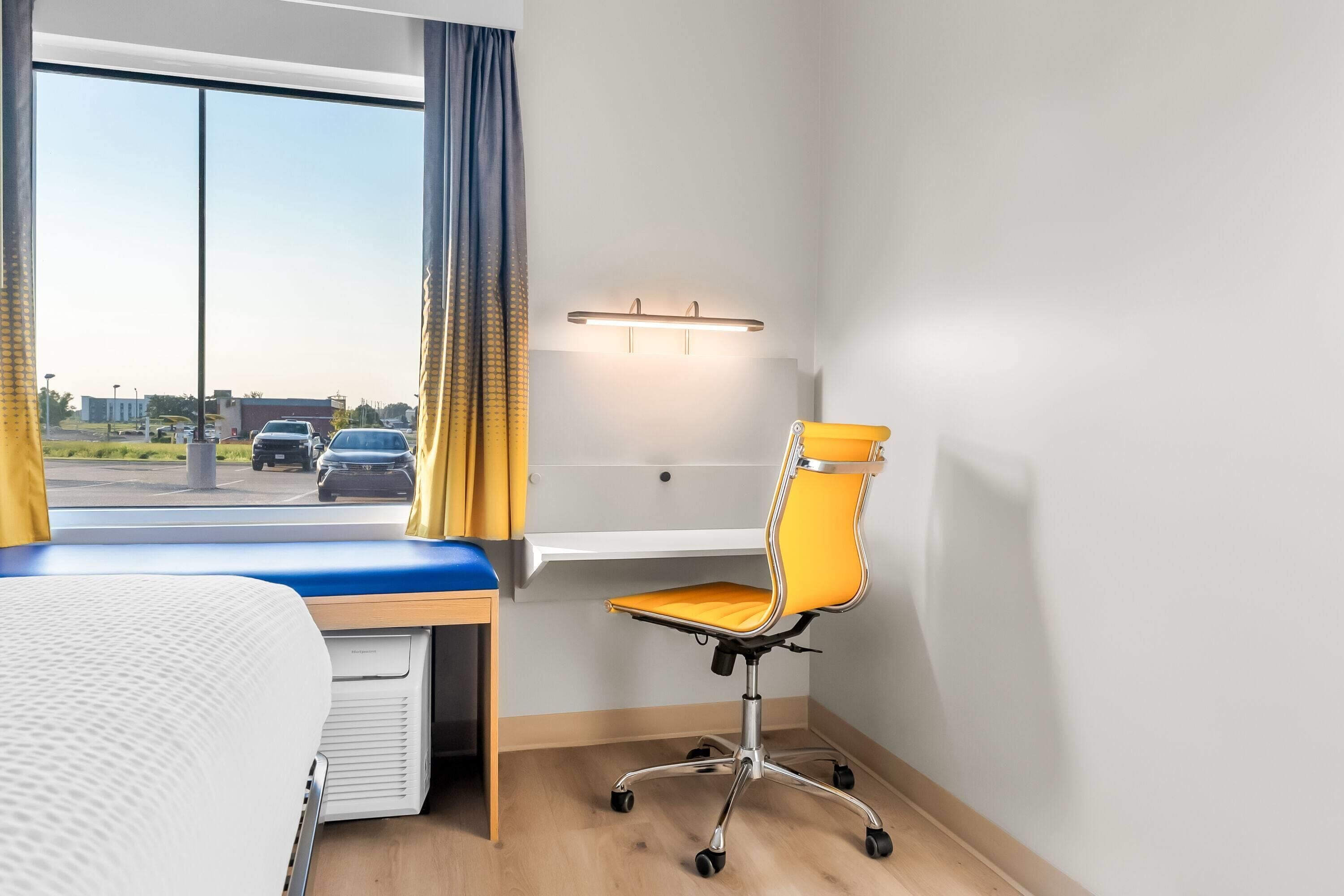 Habitación de hotel con cama, escritorio y silla, ventana con cortinas y vista al estacionamiento.