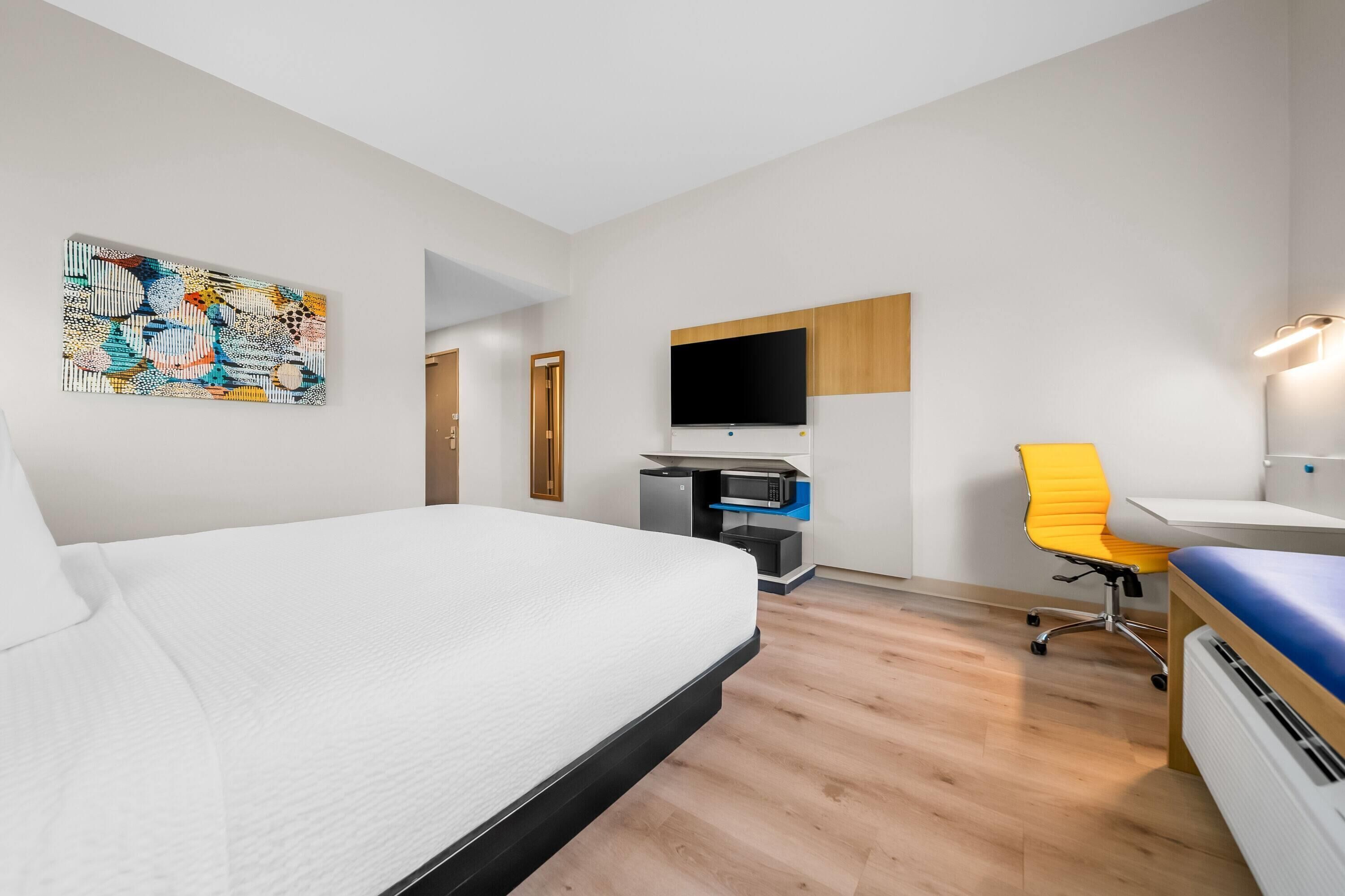 A hotel room with a bed, desk, chair, TV, and artwork on the wall.