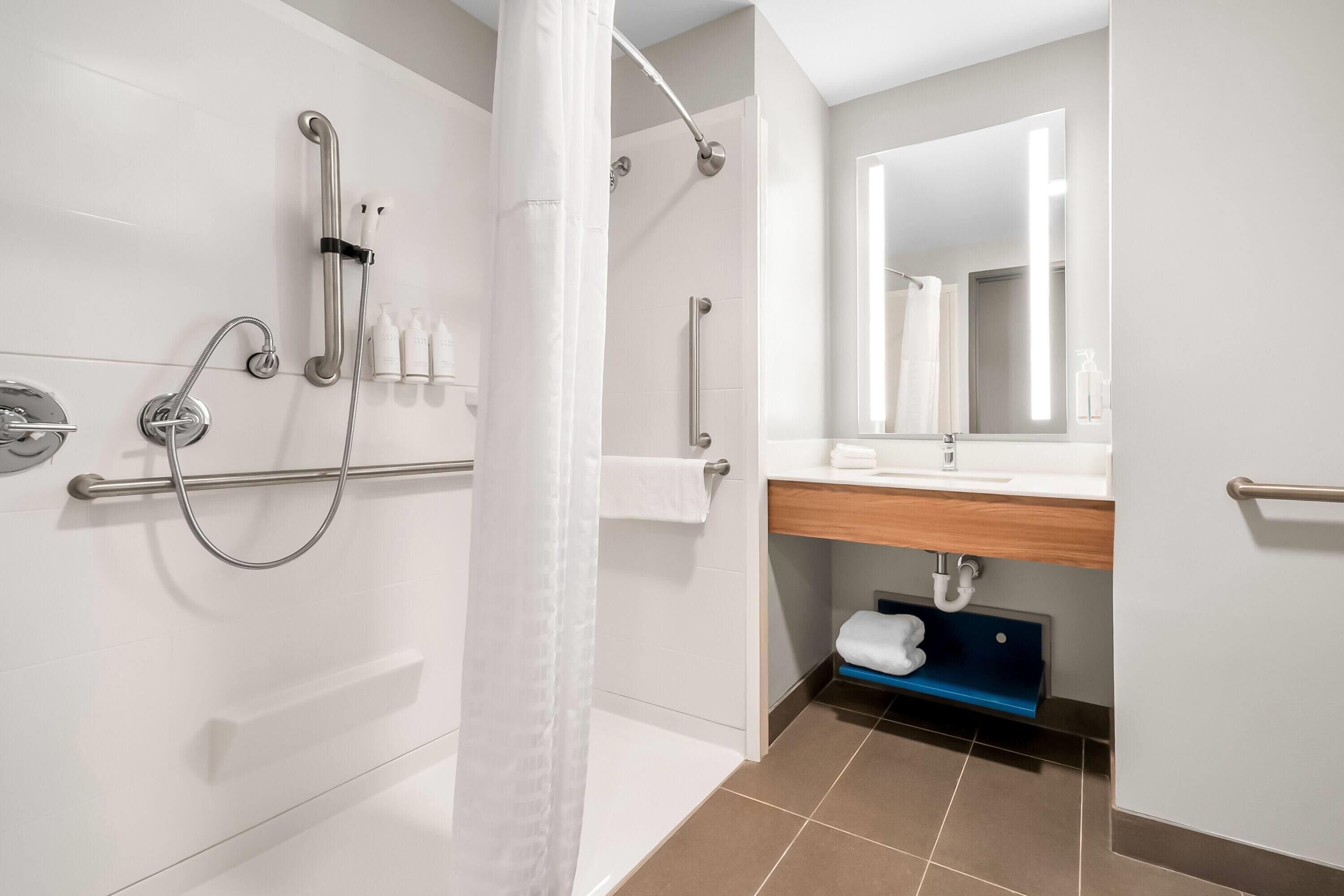 Un baño moderno con ducha a nivel, barras de apoyo, un porta papel higiénico y un espejo sobre el lavabo.