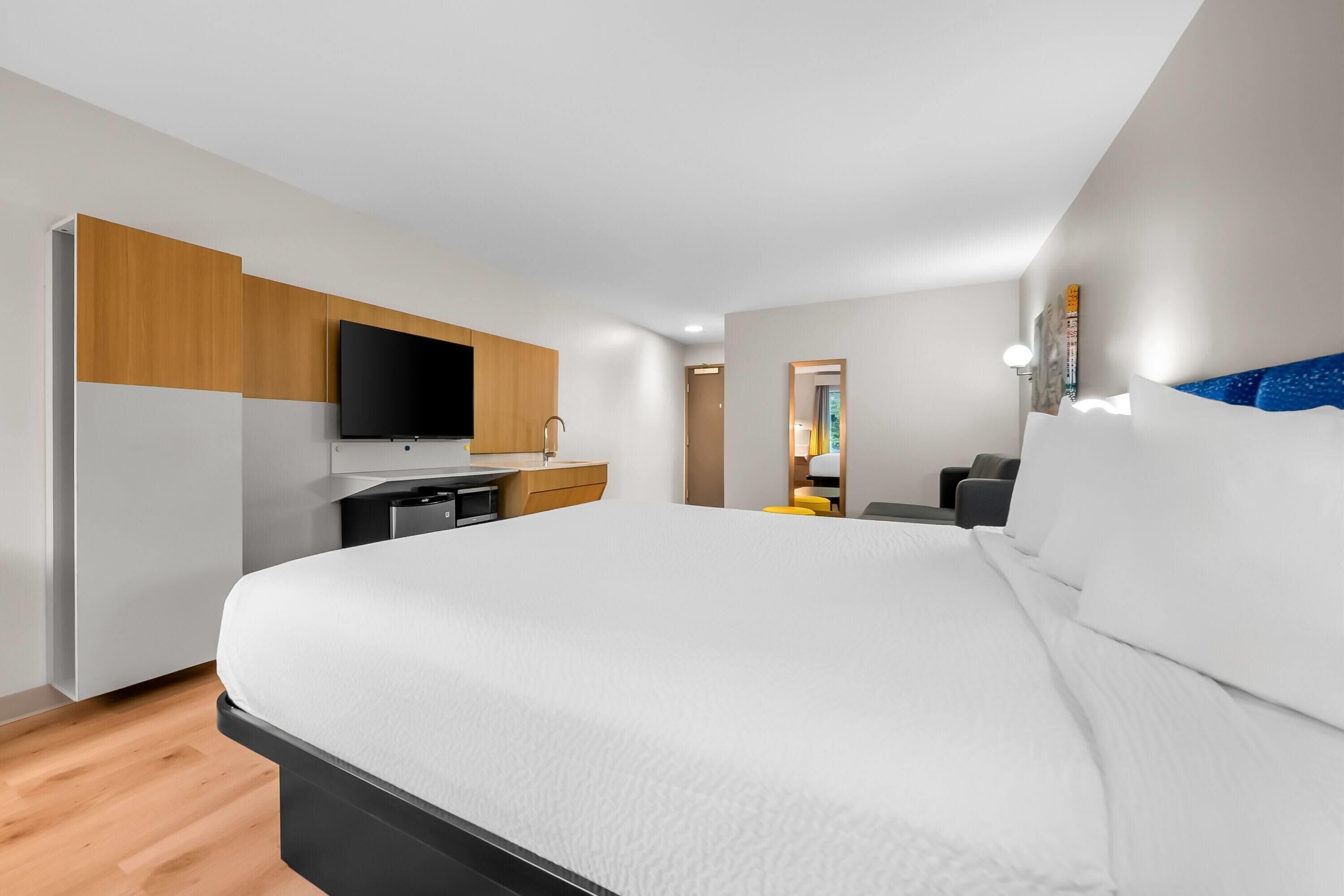 A modern hotel room with a large bed, a flat-screen TV, a wooden cabinet, and a small kitchenette.
