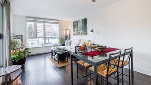 A modern living room with a glass dining table, dark wooden flooring, a ceiling fan, and large windows offering a city view. - Modern & Bright Downtown Haven ~ Pool ~ Gym ~ Pkg! (Vancouver)