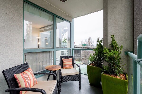 A balcony with wicker chairs and a small table, large windows, and potted plants.