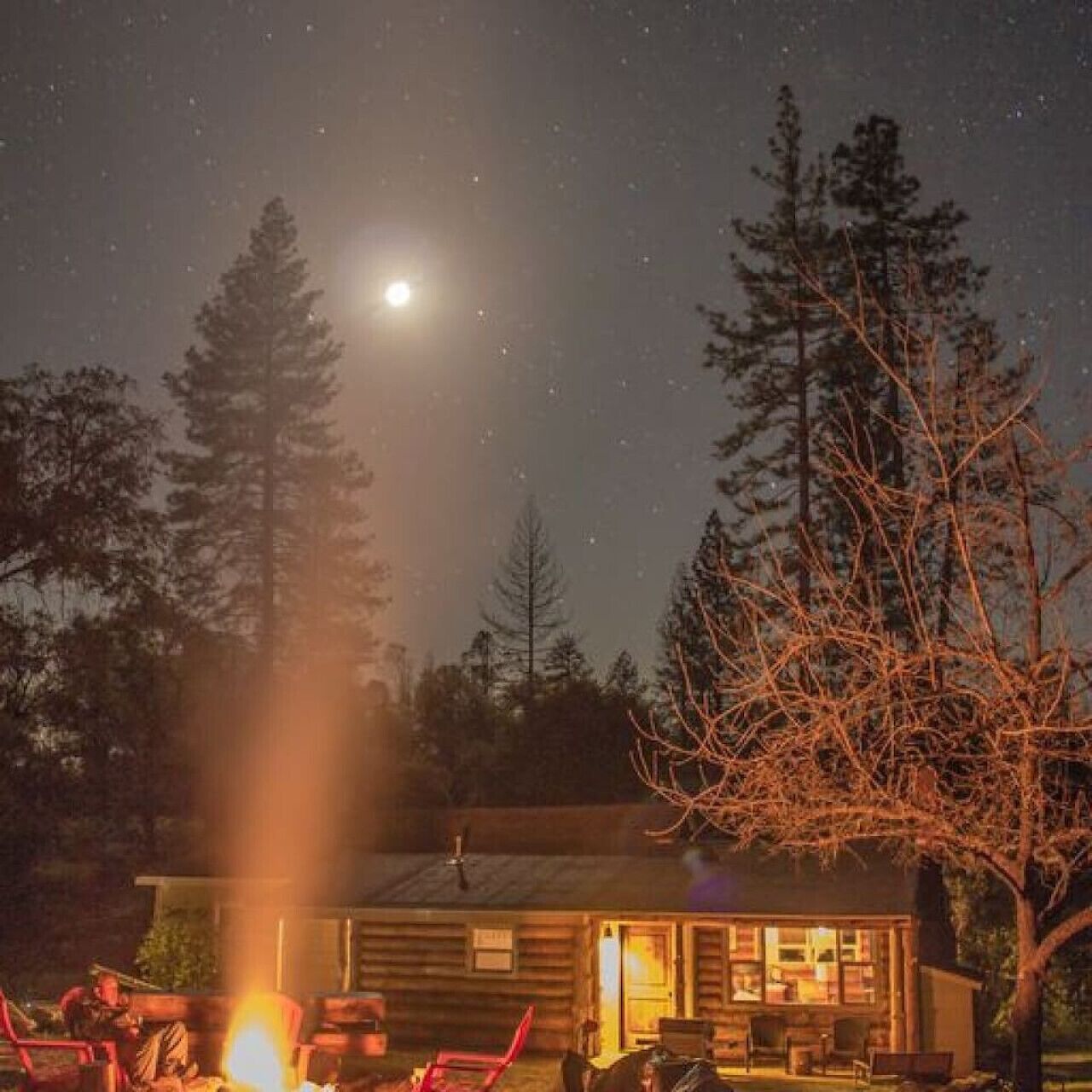 Une cabane avec une cheminée bien visible, un petit espace de véranda et un ciel nocturne dégagé parsemé d’étoiles.