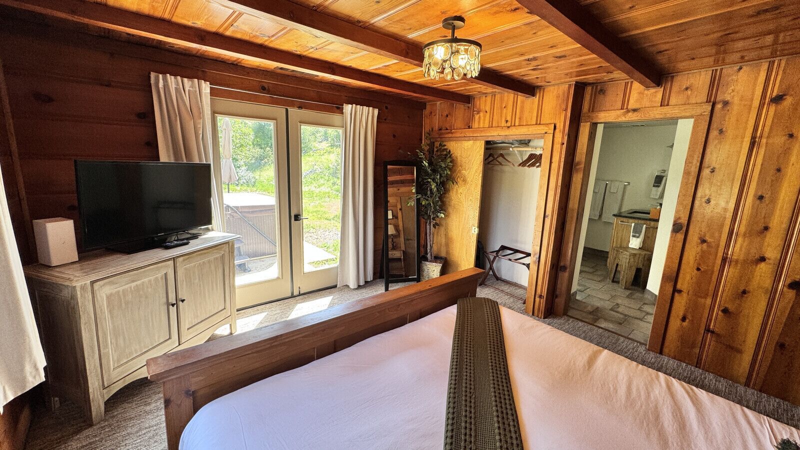 A wooden cabin bedroom with a bed, a television on a cabinet, and a sliding glass door leading to an outdoor area.