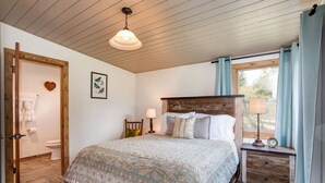 A bedroom with a wooden bed, a nightstand with a lamp, a window with curtains, and a wooden door. - Rare 12-Guest Cabins w/ Hot Tubs, Fire Pits & Pets (Mariposa)