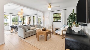 A spacious living room with a large window, a ceiling fan, and a black TV stand. - Charming Blue Mountain Bungalow in 30A. Walk or Golf Cart down to the sand.  (Santa Rosa Beach)