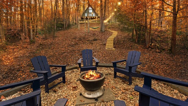 Un espace extérieur accueillant, aménagé avec un foyer et entouré de fauteuils Adirondack, qui mène à un chalet situé dans une zone boisée.