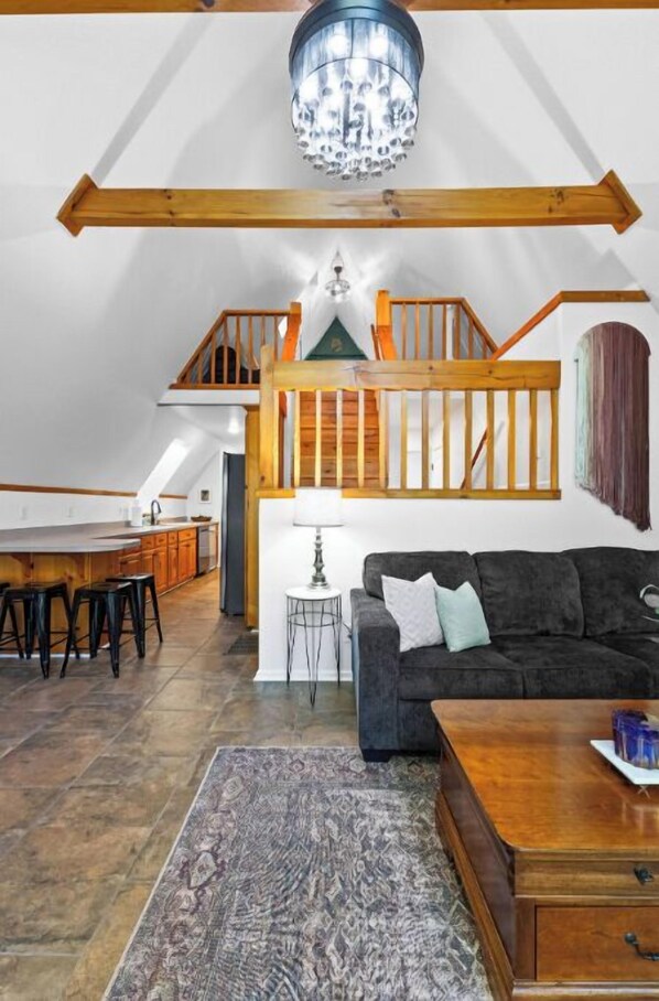 A loft-style living area with a staircase, wooden beams, a chandelier, a sofa, a coffee table, and a patterned rug.