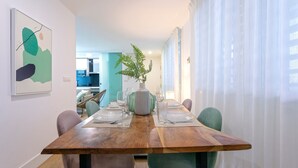 A dining area with a wooden table set for four, surrounded by chairs. A potted plant is on the table, and there is a kitchen visible in the background.