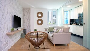 A modern living room with a sofa, a wicker chair, a flat-screen TV, and a kitchen in the background.