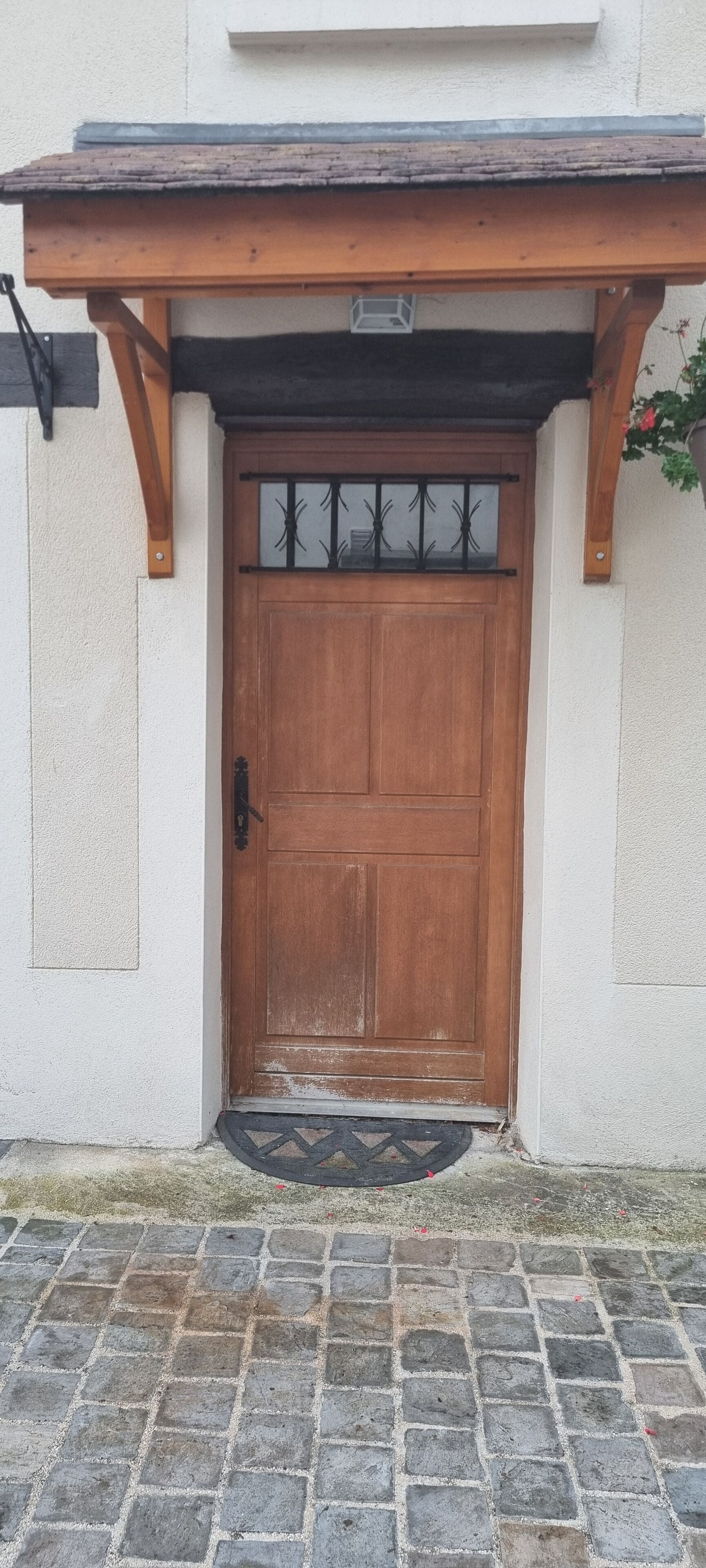 Une porte en bois avec un portail en métal, une entrée pavée de pierres et un petit porche doté d’une poutre en bois et d’une rambarde en métal.