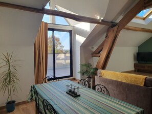Ein gemütliches Zimmer mit Esstisch, Stühlen, einer Couch, einem Fernseher und einem großen Fenster mit Vorhängen.