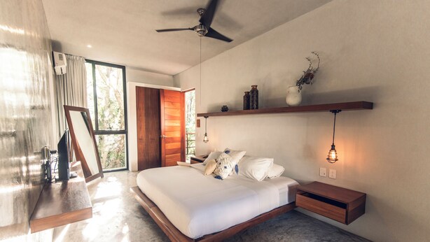 Un dormitorio moderno con una cama grande, un ventilador de techo, una mesita de noche de madera, una mesilla auxiliar, un estante en la pared y una lámpara colgante.