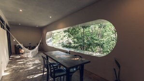 Una habitación con un amplio ventanal circular que ofrece vistas a un bosque. Hay una mesa de madera con sillas, una hamaca y una pequeña escalera.