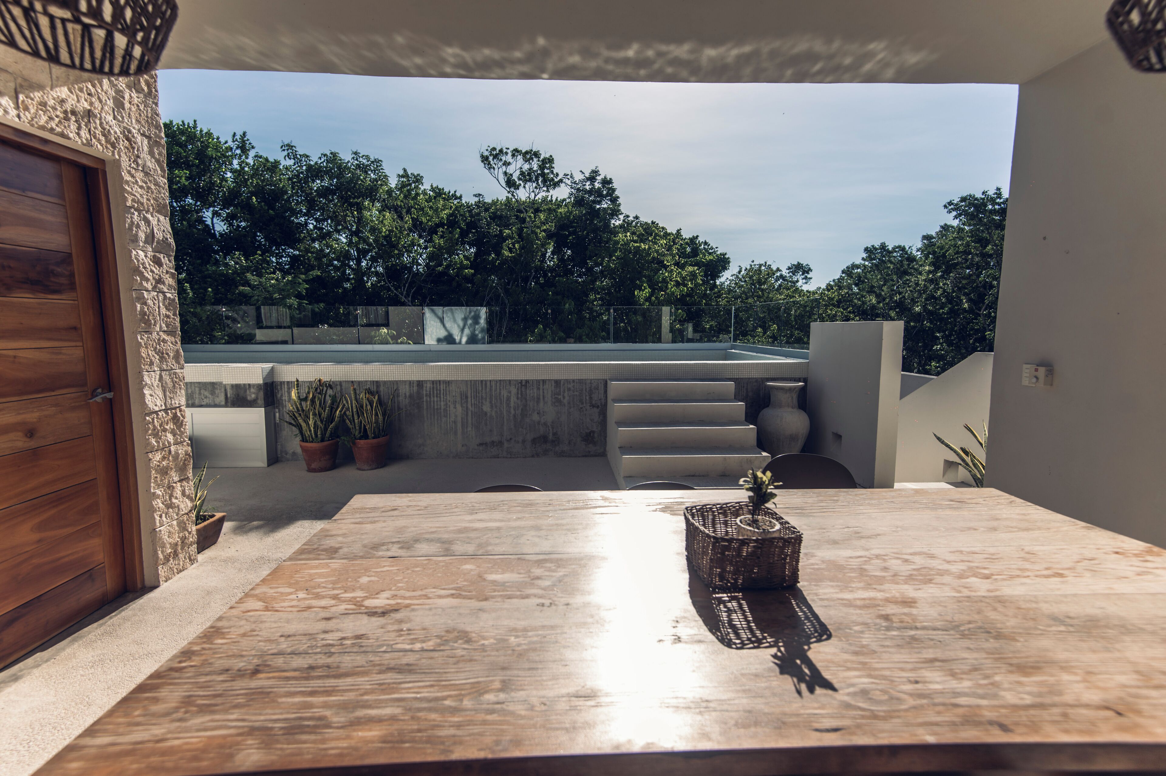 Un patio con mesa de madera, plantas en macetas y vistas a una piscina y vegetación.