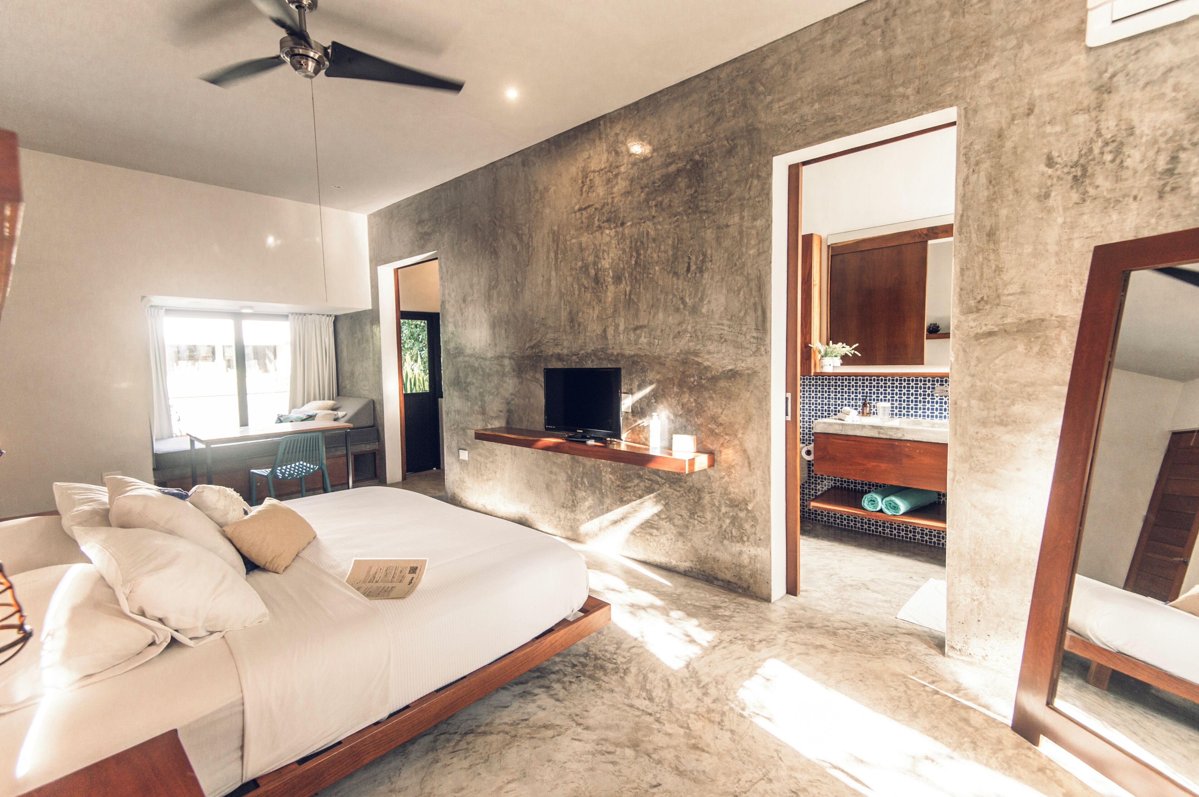 A modern bedroom with a bed, a TV on a shelf, a desk, and a bathroom with a mirror and sink.