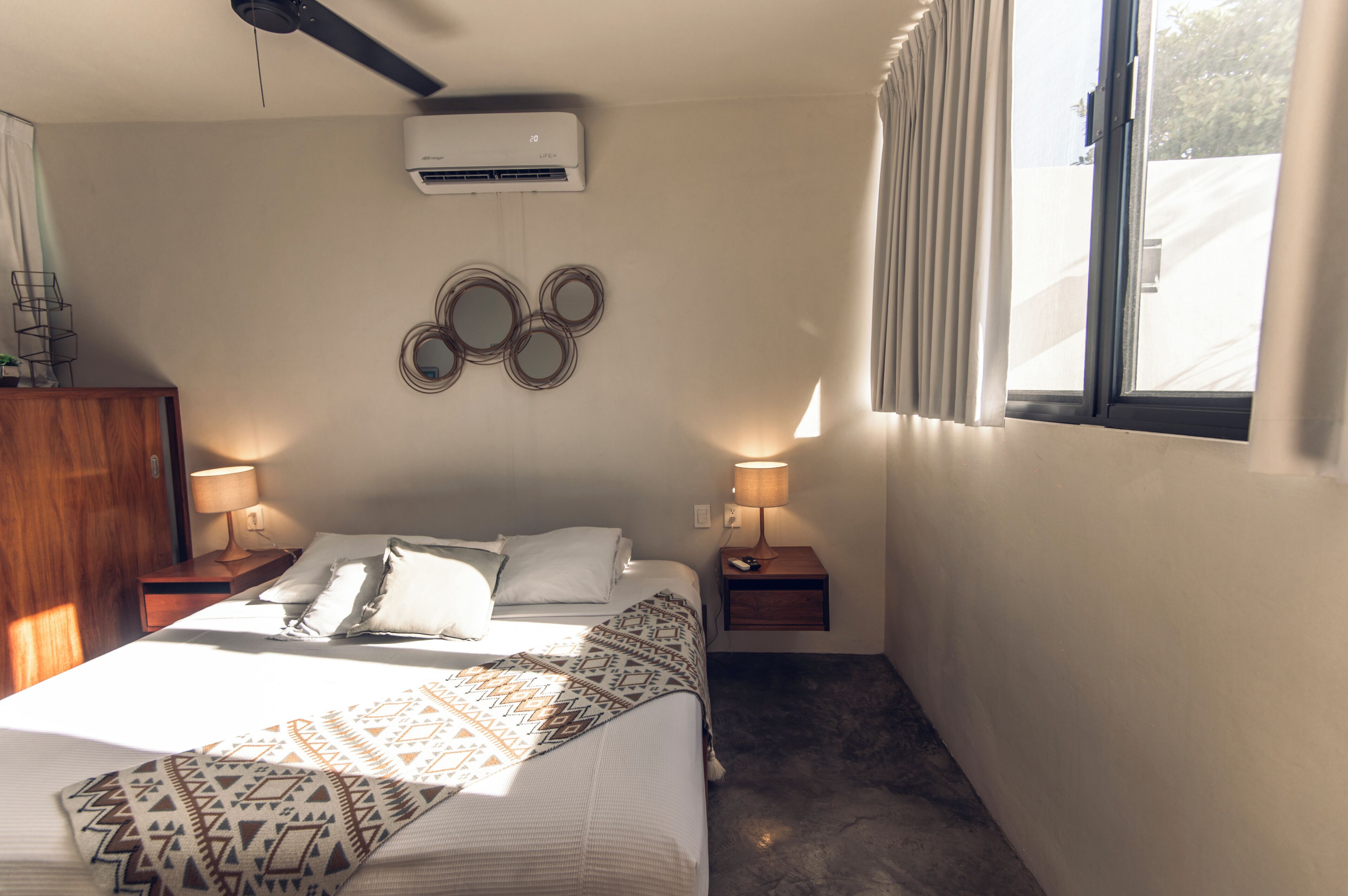 A hotel room with a bed, two bedside tables with lamps, an air conditioning unit, a window with curtains, and a wall decoration of circular mirrors.
