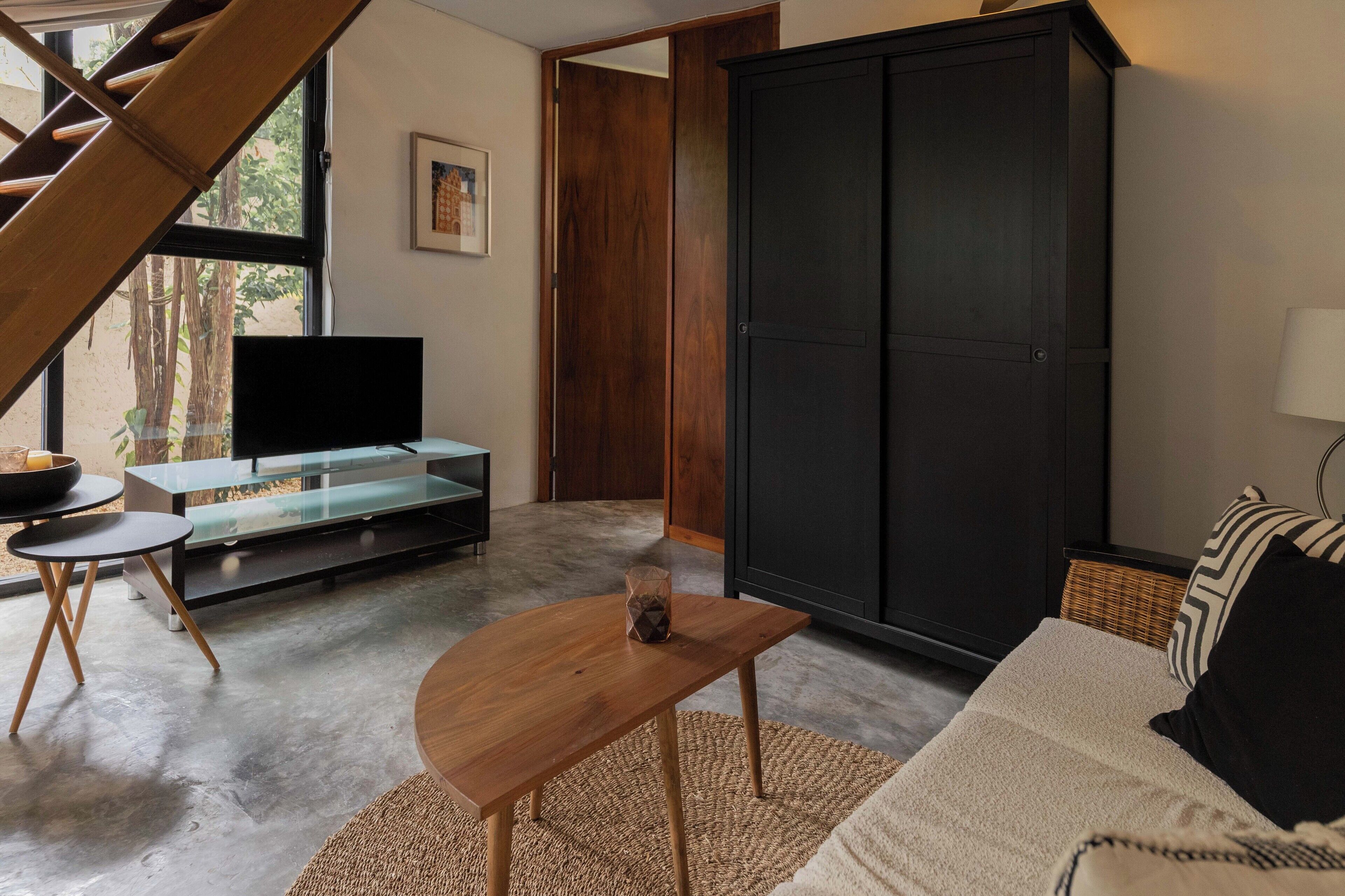 A modern living room with a wooden coffee table, a glass-top TV stand, a dark wardrobe, and a sofa with patterned cushions.