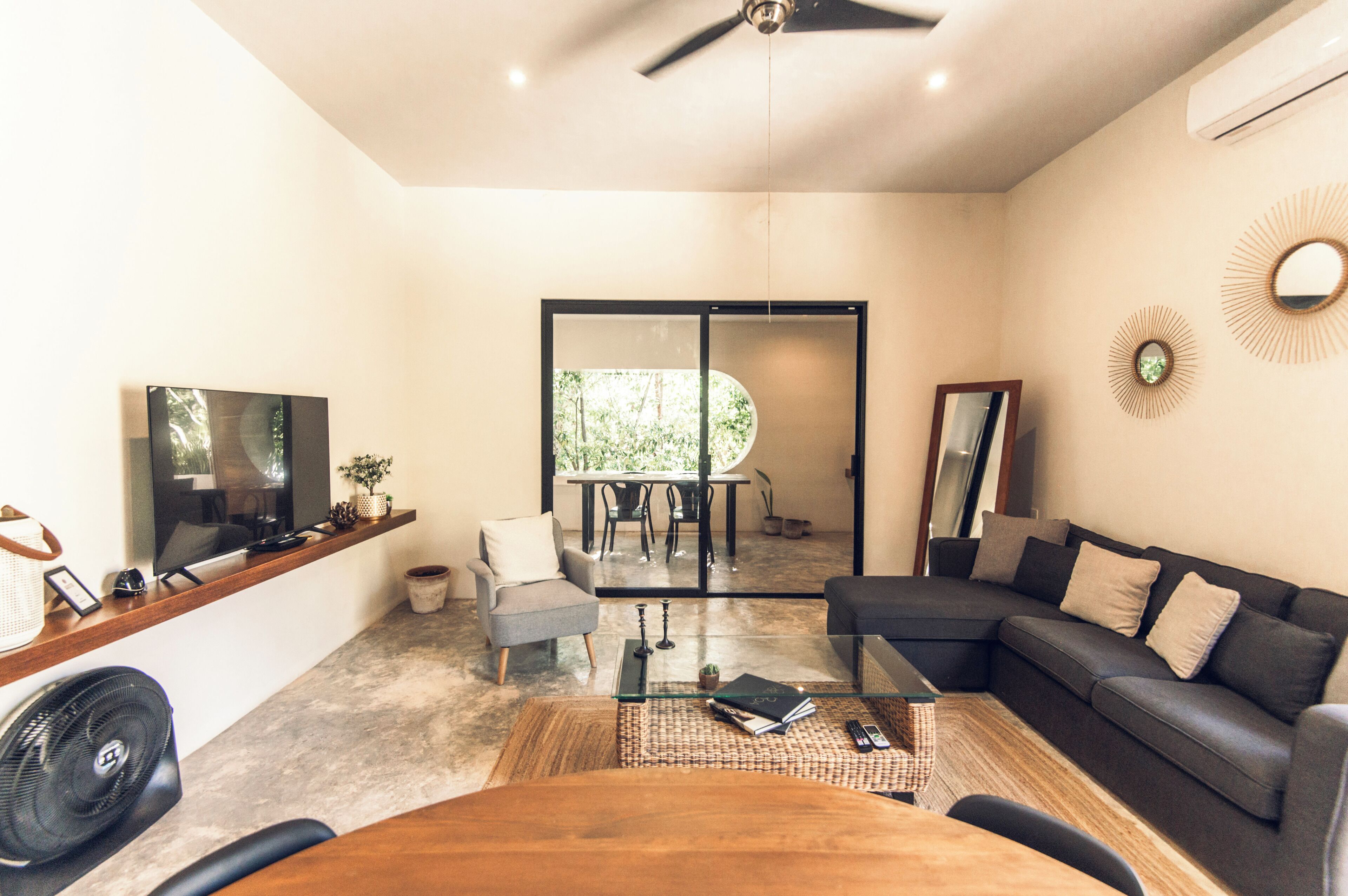 Un salon moderne avec un canapé d’angle, une table basse en verre, un téléviseur posé sur une étagère en bois et un ventilateur de plafond.