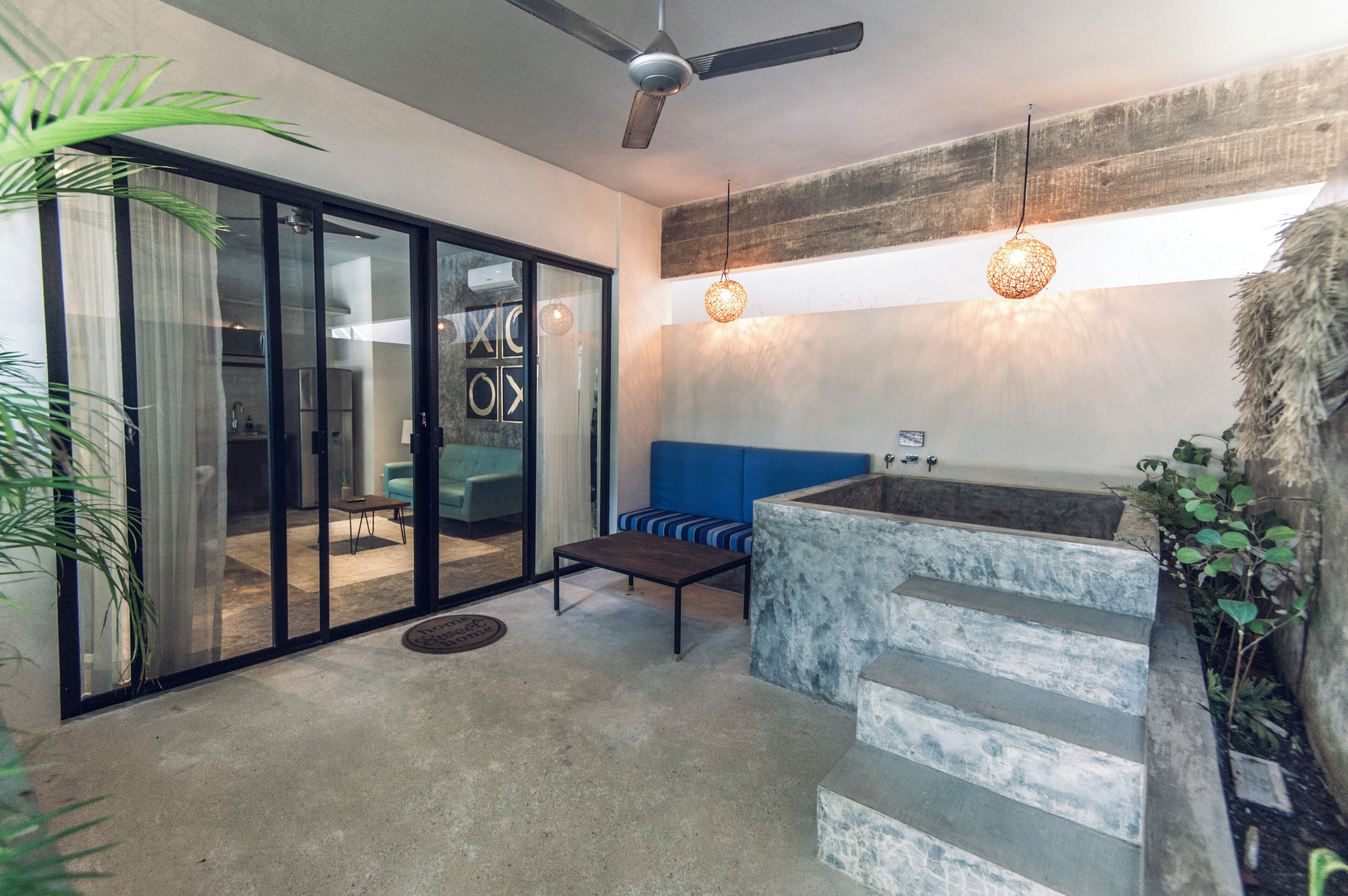 A modern interior with a glass door leading to a kitchen, a blue cushioned bench, a concrete water feature, and a ceiling fan.