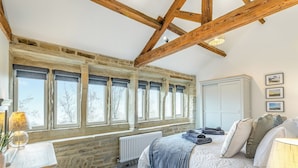 Un dormitorio con pared de piedra, vigas de madera, una cama con manta azul, un radiador y una ventana con vistas a árboles.