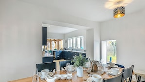 Un comedor con mesa de madera dispuesta para seis comensales, una cocina al fondo y una lámpara colgante moderna.