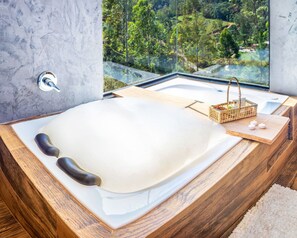 A modern bathroom with a large bathtub, a wooden vanity, and a view of a lush landscape through a large window. - Vila nas Montanhas (Anitapolis)
