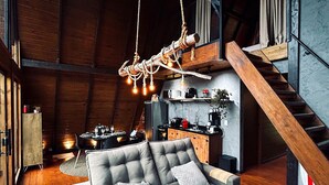 Moderni loft-tyylinen asunto, jossa on esillä puupalkit, harmaa sohva koristetyynyineen, riippuvalaisin ja portaikko.