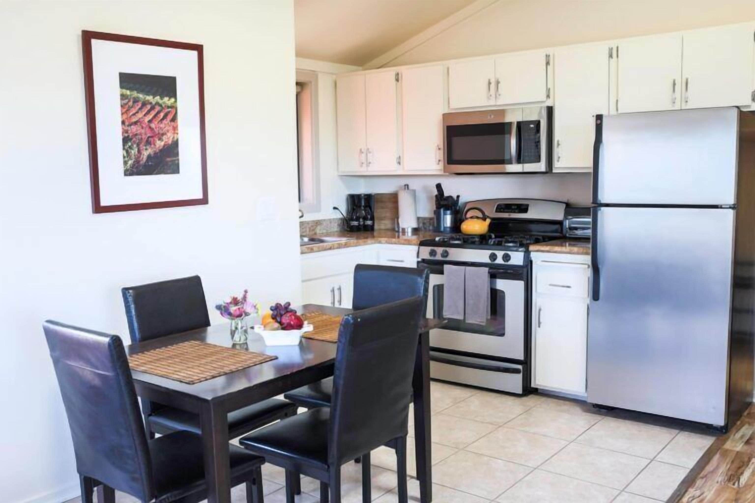 A kitchen with white cabinets, a stainless steel refrigerator, a microwave, and a stove. There is a dining table with chairs and a vase with flowers.