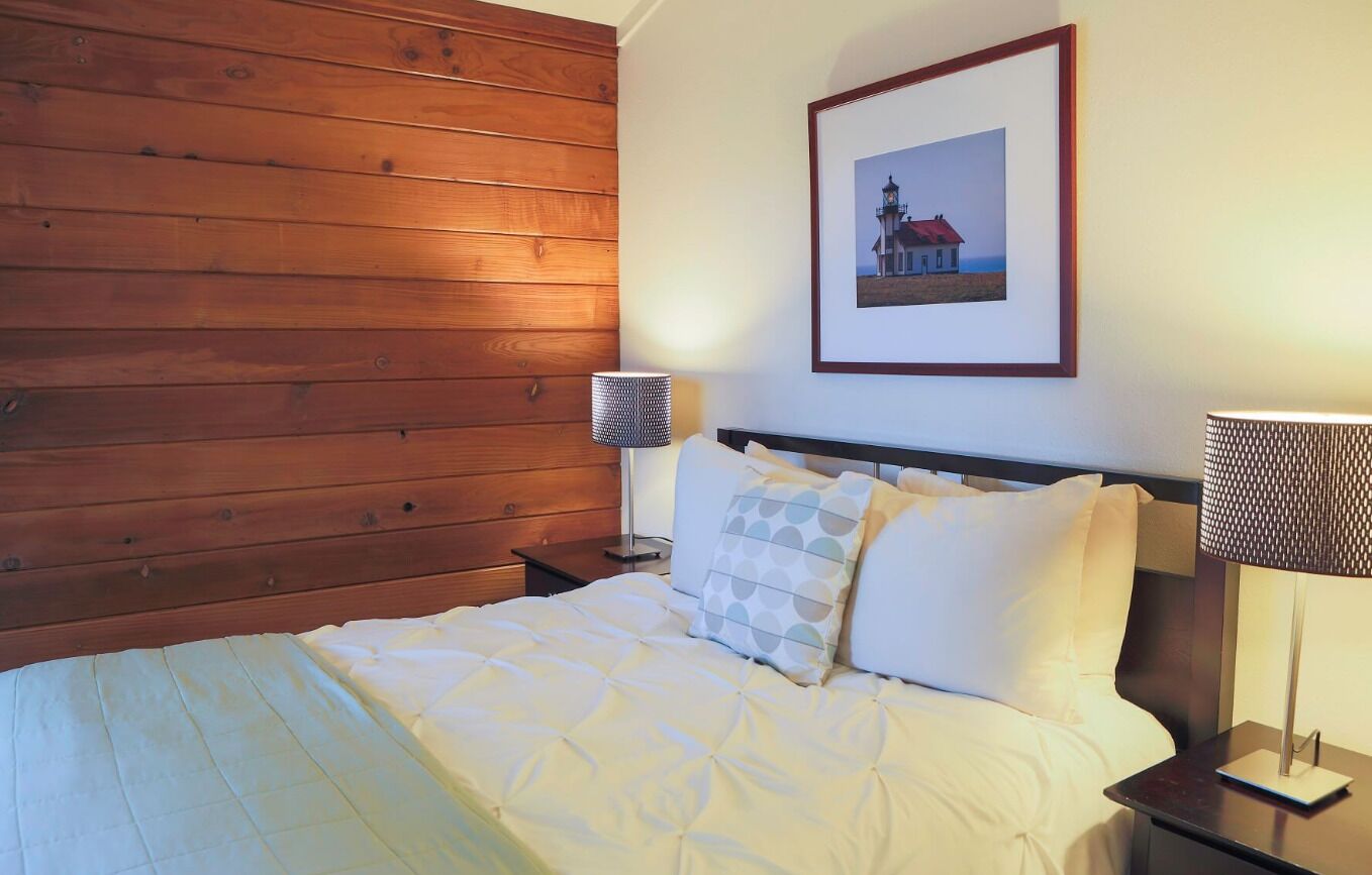 A neatly made bed with a white duvet and patterned pillows, a framed picture of a lighthouse on the wall, and two bedside lamps on wooden nightstands.