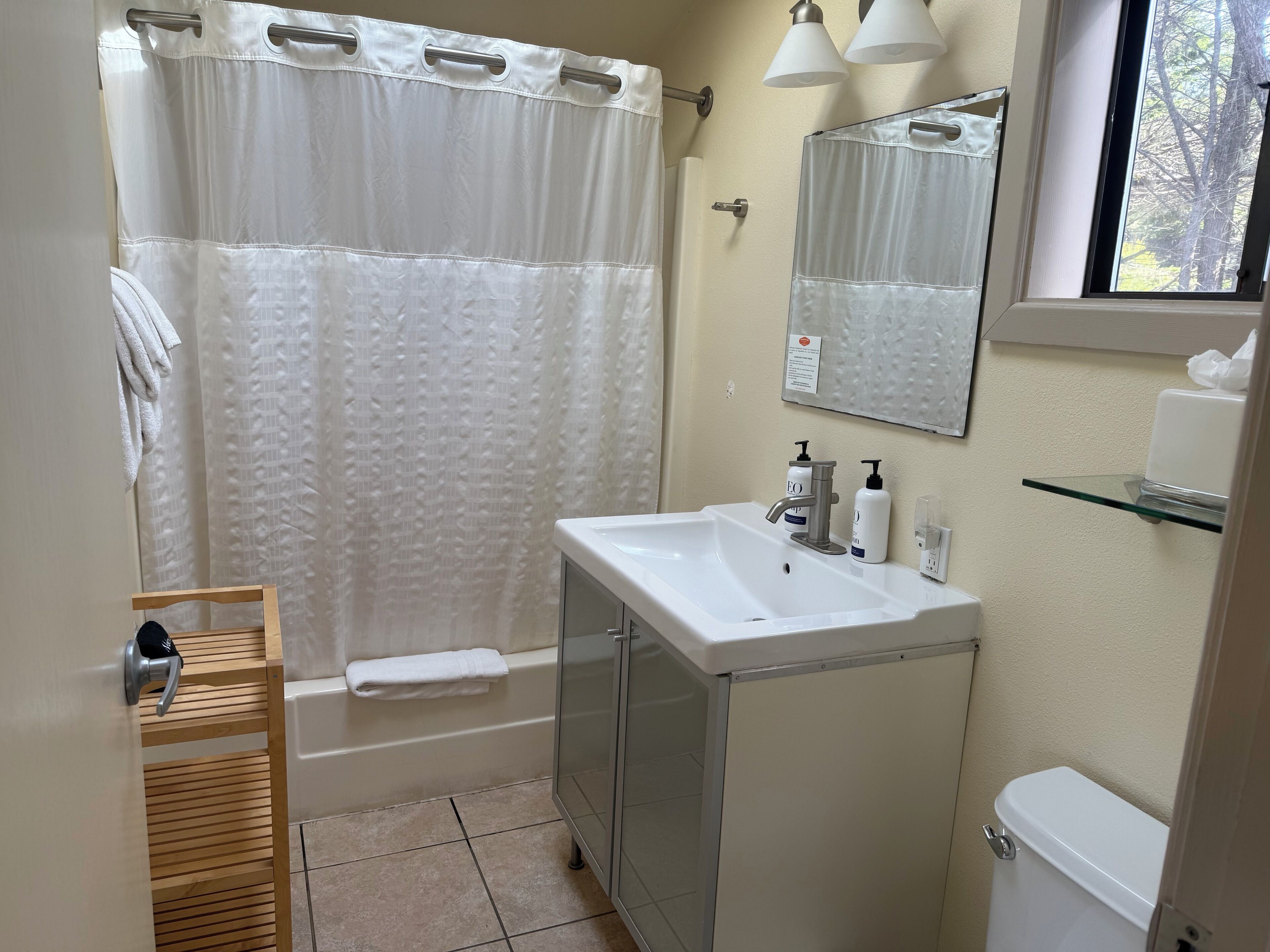 A bathroom with a shower curtain, a sink with toiletries, a glass shelf, and a toilet.