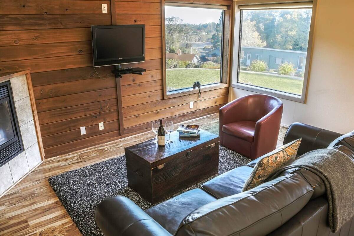 Una sala de estar con pared de madera, televisor, chimenea, sofá de cuero, sillón rojo y una mesa de centro con una botella y un libro.