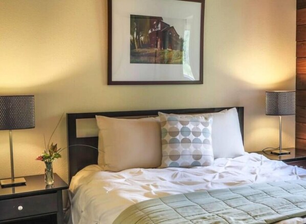 A neatly made bed with a quilted blanket, two bedside tables with lamps, a framed picture on the wall, and a vase with flowers on the nightstand. - Cottages at Little River Cove (Little River)