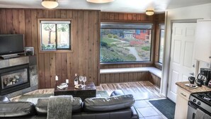 Una sala de estar acogedora con chimenea, sofá de cuero y un amplio ventanal con vistas a un jardín y un edificio rojo.