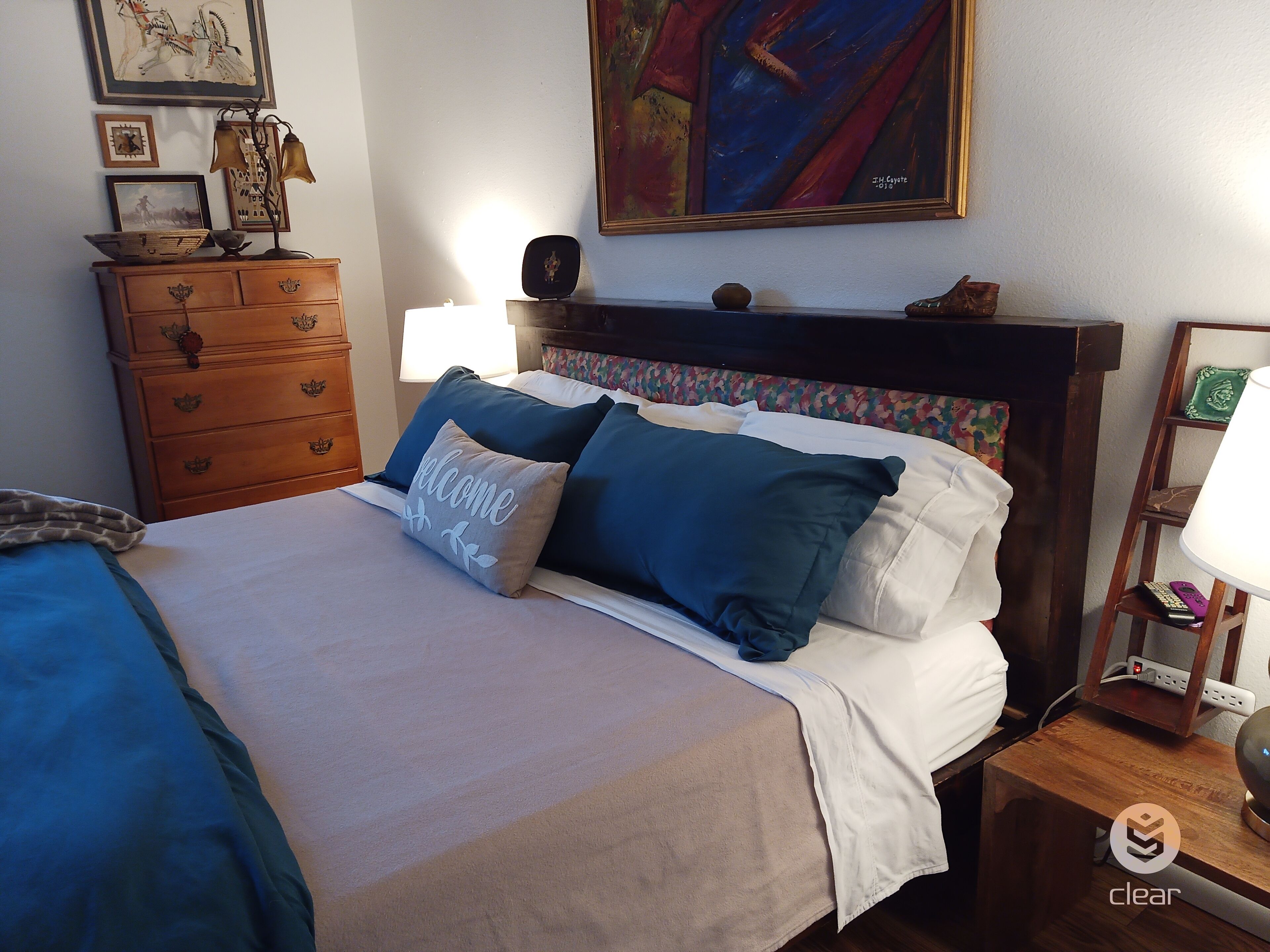 A neatly made bed with blue and white bedding, a wooden dresser with drawers, a bedside table with a lamp, and a painting on the wall.