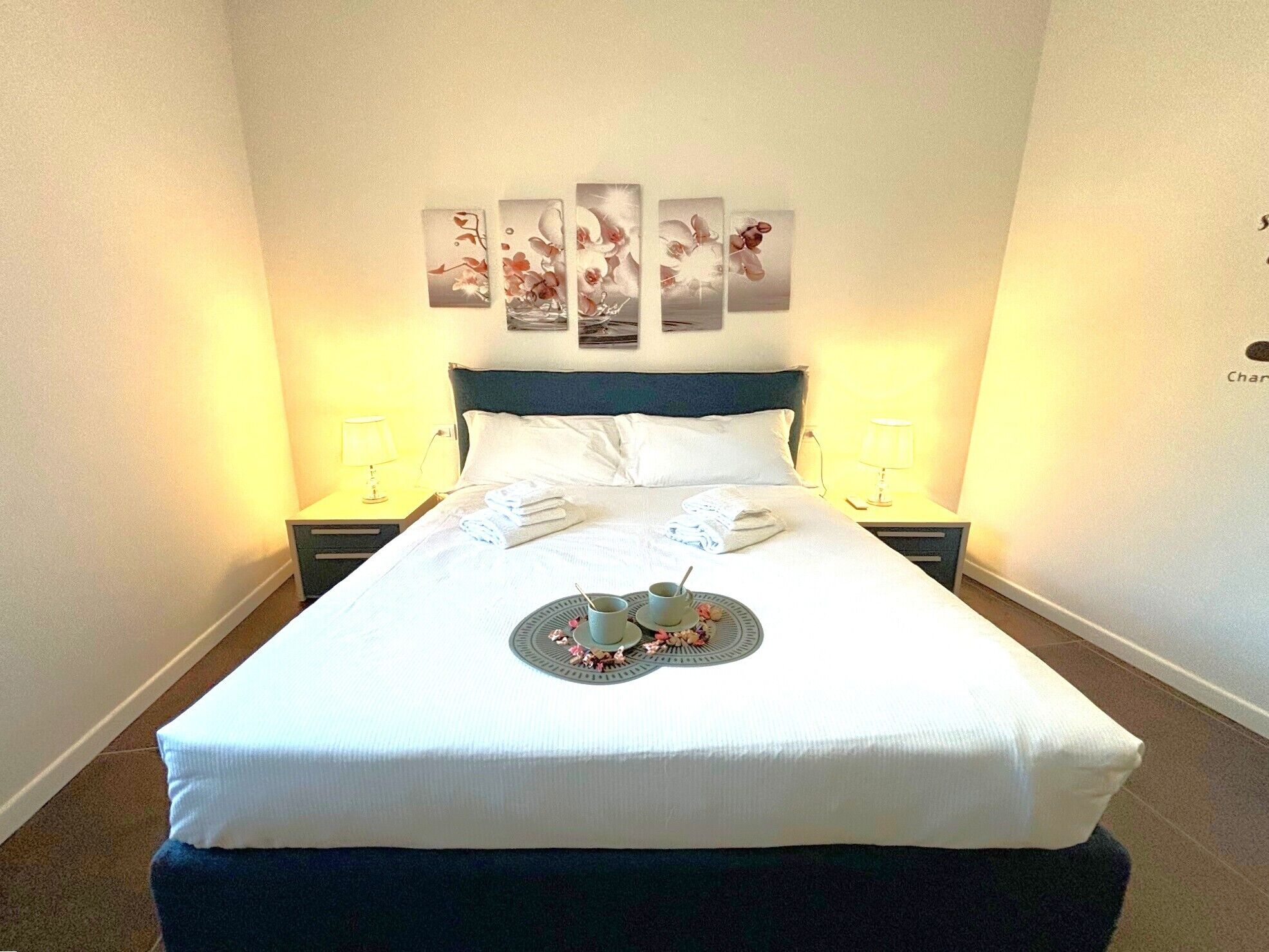 Cama bem arrumada com toalhas dobradas, bandeja com xícara e flores, e arte nas paredes acima.