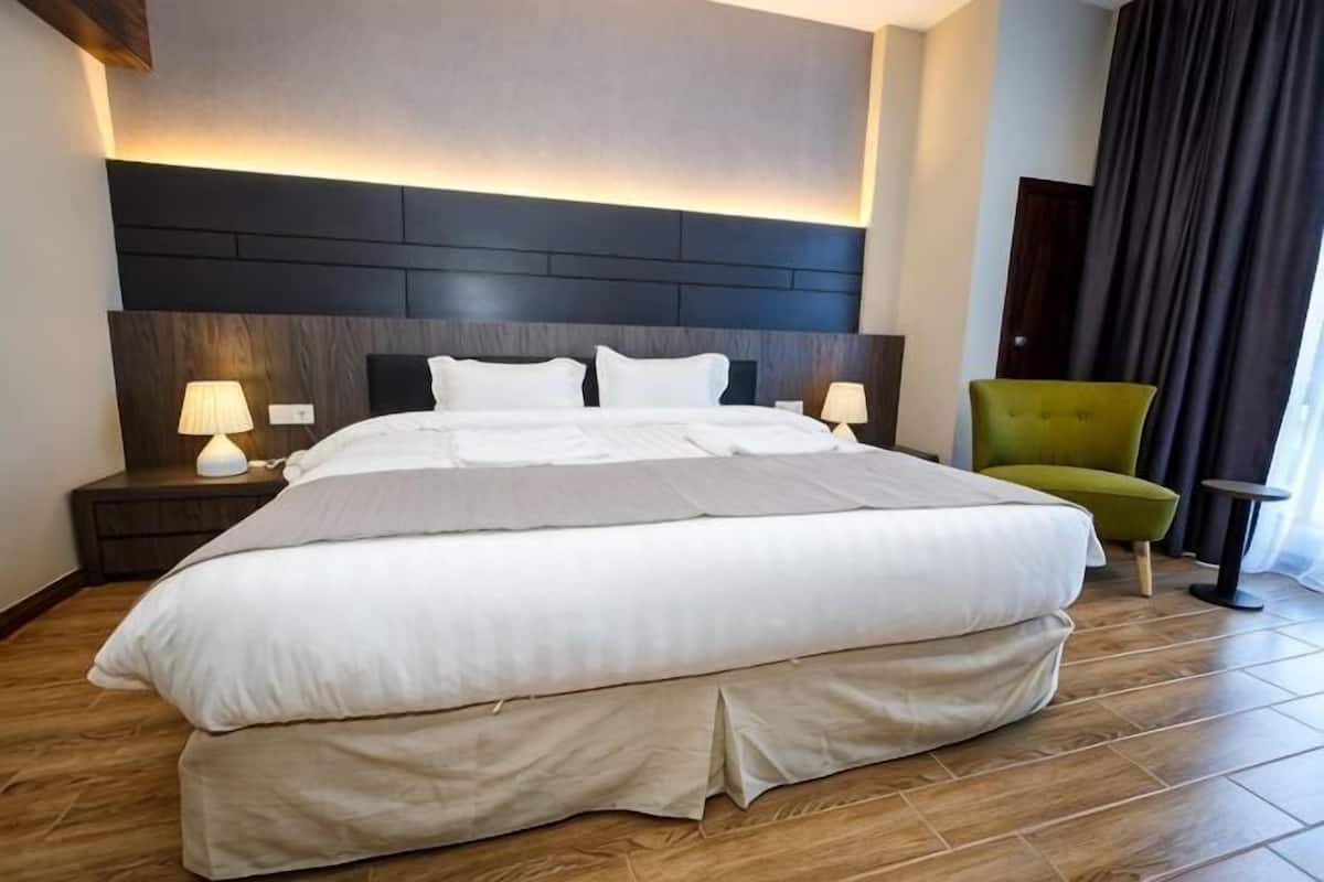 A hotel room with a large bed, two bedside tables with lamps, a green armchair, and a dark headboard.