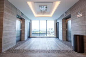 Un hall d’ascenseur moderne avec vue sur la ville, deux ascenseurs et un panneau indiquant qu’il est interdit de fumer.