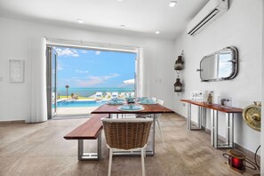 A modern dining area with a wooden table and chairs, a wicker basket, and a view of the ocean through large glass doors. - VILLA JOYUDA (Cabo Rojo)