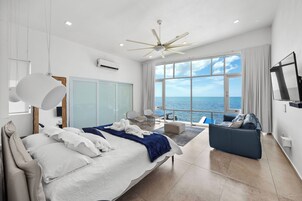 Un dormitorio moderno con cama, sofá, ventilador de techo y una ventana grande con vista al mar.
